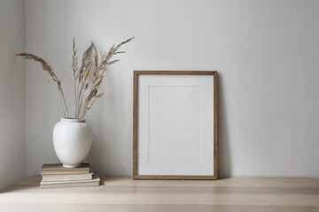White ceramic vase with dry flowers, grass on old books. Blank vertical wooden picture frame...