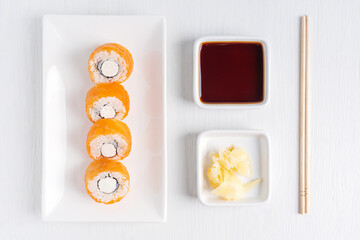 Top view of portion of traditional asian sushi roll made with raw or smoked salmon fish, boiled rice, cream cheese and nori seaweed served on plate with ginger, soy sauce and chopsticks on white table