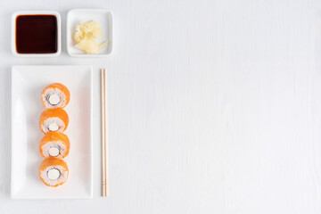 Top view of asian sushi roll made with raw salmon fish, boiled rice, cream cheese and nori seaweed served on plate with ginger, soy sauce and chopsticks on white wooden background with copy space