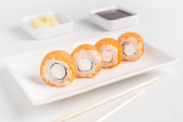 Portion of fresh healthy asian sushi roll made with raw or smoked salmon fish, cream cheese, nori seaweed and boiled rice served on plate with ginger, soy sauce and chopsticks on white wooden table