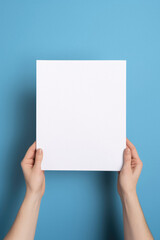 Hands holding blank paper sheet on blue background. Mockup for design.