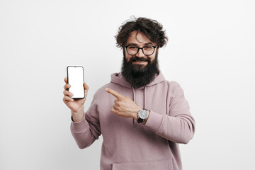 Joyful bearded man in a pink hoodie pointing at a blank smartphone screen, ideal for app promotion, isolated on a white background