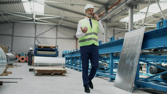 Caucasian inspector with reflection vest and safety helmet walking along metal technical machine in warehouse. Busy man talking on his smartphone. In background workers producing industrial goods.