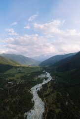 Fototapeta premium Vertical photo, The valley and the town of Mazeri in the distance. Aerial view of the stormy flow of the Dolra River, in the middle of a forest, a hiking trail, a tourist route. Svaneti, Georgia