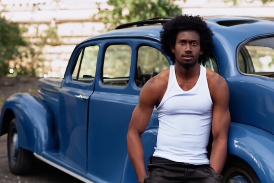 a guy stands near a retro car. a young afro guy with curly hair in a white T-shirt and black pants stands near a blue retro car
