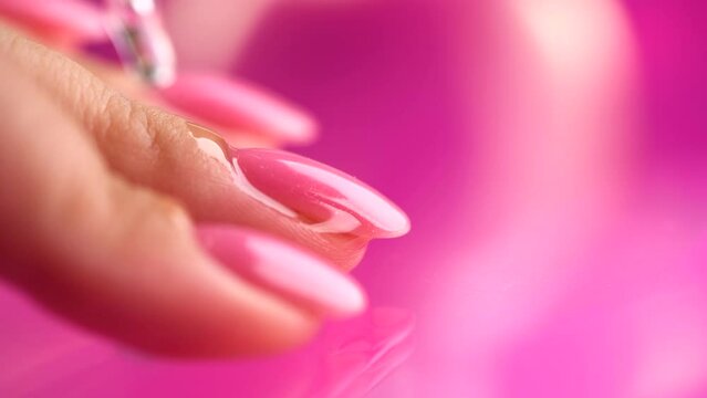 Applying cuticle oil in pipette, on fingers, manicure salon. Nail care, polish, pink shellac UV gel, varnish, manicure process in beauty salon. Over pink background. Application of nails oil closeup