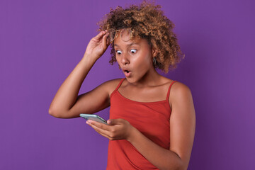 Young shocked ethnic African American woman teen lifts glasses from eyes seeing advertisement on phone with very advantageous shopping offer or shocking prices stands in purple studio.