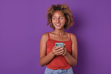 Young happy thoughtful African American woman blogger holding phone coming up with new post for social network wanting to surprise subscribers or talk about past weekend stands on purple background. - obrazy, fototapety, plakaty