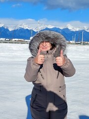 A woman in winter clothes stands in the snow against the background of snow-capped mountains.