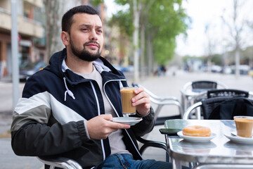 Young bearded man traveler enjoying warm cocoa with sweet soft donut at table of street cafe while...