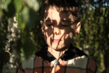 Boy basks in dappled light, a peaceful expression on his face. The image aligns with wellness and mental health awareness, illustrating the calming effect of nature.