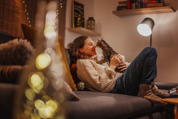 Cozy at home with tabby cat, woman with her pet on sofa ay home in evening, winter holidays concept - obrazy, fototapety, plakaty