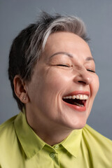 Obraz na płótnie Canvas cheerful funny 50s elderly woman with short gray hair laughing loudly on grey background