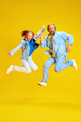 Father and daughter, parent and kid dressed style denim outfit and have fun listening music in headphones and jumping of joy isolated yellow background.