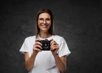 Cheerful woman in glasses and a white t-shirt holding a camera against a dark background