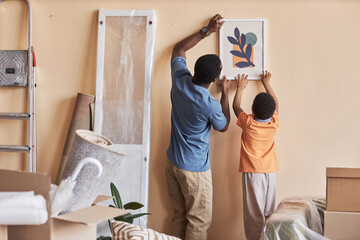 Rear view of African American man and his little son hanging picture on wall of spacious living room during relocation to new apartment