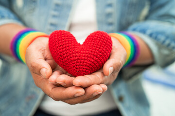 Asian lady wearing rainbow flag wristbands and hold red heart, symbol of LGBT pride month celebrate annual in June social of gay, lesbian, bisexual, transgender, human rights. - obrazy, fototapety, plakaty