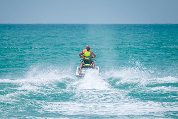 Two persons wearing safety life jackets drive jetski from shore to open sea, splashing water....