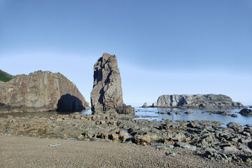 A rocky beach with a tall, dark basalt formation in the center, surrounded by smaller rocks under a...