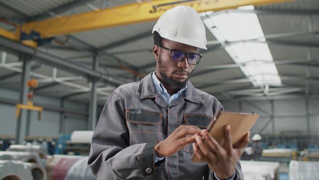 Attractive African American man remotely chatting on tablet device with customer while standing in large industrial workshop. Using digital device while working in manufacturing plant.