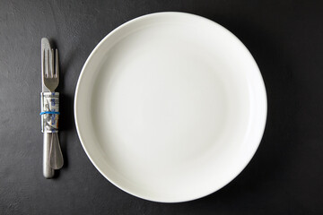 Empty white ceramic plate with cutlery wrapped in hundred dollar bill on black stone table. Top...