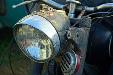 glass round large front powerful old iron chrome shiny classic electric halogen headlight with an iron steering wheel from an old retro black motorcycle on the street during the day