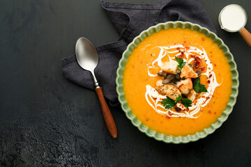 Pumpkin and carrot cream soup with herbs, seasonings and seeds in bowl on black wooden background in rustic style. Thanksgiving traditional autumn pumpkin cream soup. Top view.