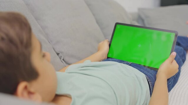 Close up kid lying on sofa, holding and tapping on a tablet with green screen. 