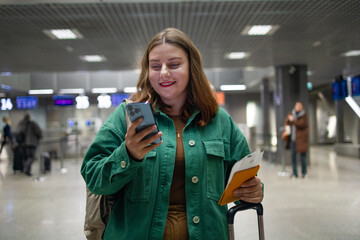 Smiling woman walking with luggage in airport terminal, cheerful female holding passport with tickets and carrying suitcase during departure departure