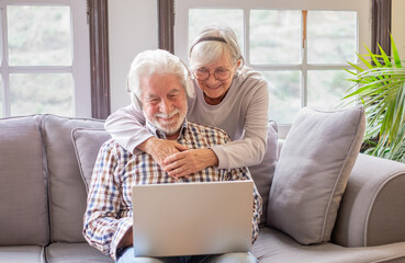 Happy 70s white-haired senior couple relaxing on home couch, looking in internet store using...