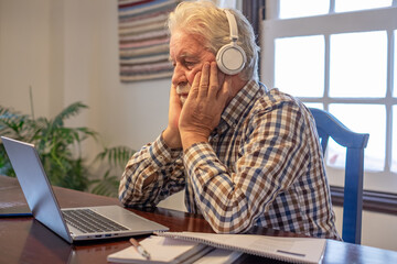 Concentrated mature senior bearded man wearing headphones sitting at table with laptop and books...
