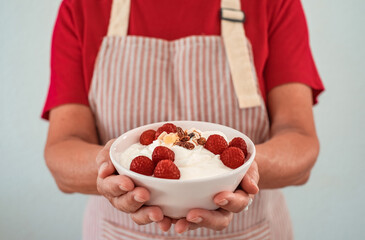 Elderly woman in apron holding bowl of yogurt filled with fresh raspberries and dried fruit....