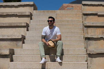 Handsome young man dressed in casual clothes and sunglasses sitting on the stairs of the park. The man has a hat and enjoys the sunny day in spring. Travel and holiday concept.