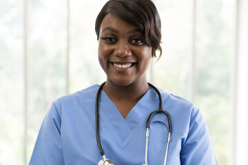 Closeup of happy and smiling African American young female doctor wearing blue scrubs uniform and stethoscope and standing at hospital
