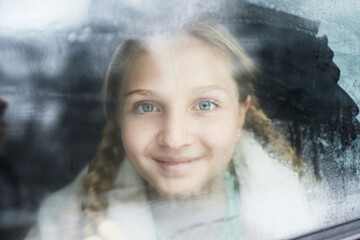 Cute smiling pre-teen girl with pigtails looking at camera while sitting in car behind steamy window and enjoying family travel