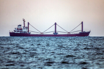 A long red ship with tall cranes on board sails in the ocean on the horizon