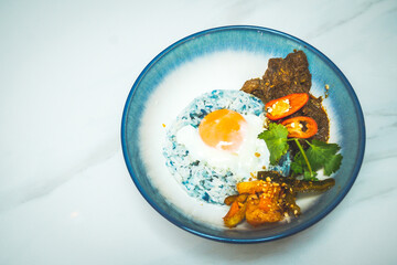 a bowl of beef rendang rice with beef rendang cuves, butterfly pea rice, sunny side up egg and pickled archar