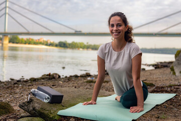 Fototapeta na wymiar A Caucasian girl on a mat by the river, doing stretching exercises to warm up before her yoga class.