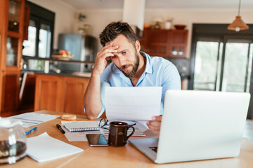 Young man holding document,paperwork,bill looking stressed at home - obrazy, fototapety, plakaty
