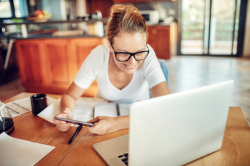 Smiling young woman using laptop with paperwork on desk at home