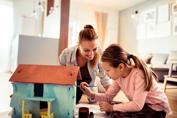 Little girl painting model house for school project with mother at home