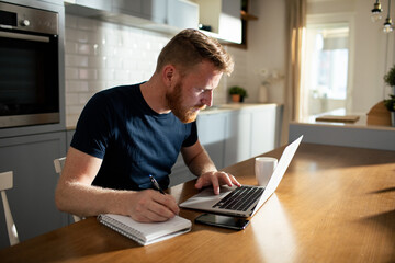 Bearded man using laptop taking notes at home - obrazy, fototapety, plakaty
