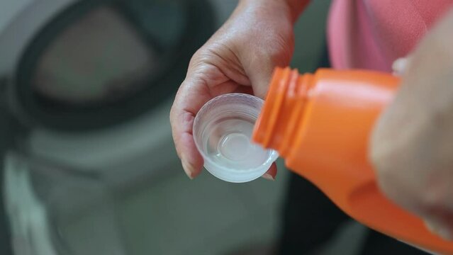 Woman pours liquid washing gel into plastic cap against background of drum of steel-colored washing machine. A girl carefully pours a transparent conditioner for flattening laundry. High quality