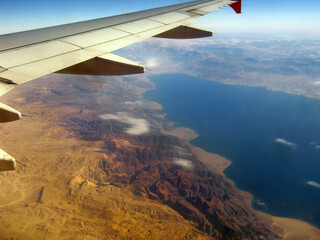 Gray airplane wing in the sky with a view of the mountains, a large lake and clouds