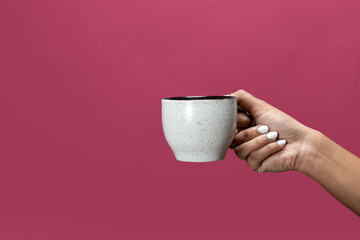 Close up on female hand, holding cup of coffee or espresso against dark pink background