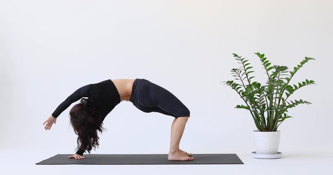 Yoga woman performs the Urdhva Dhanurasana exercise, bow pose, training in sportswear, breeches and a short top with long sleeves while sitting on a mat in the studio on a light background