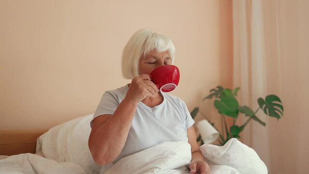 Cheerful 60s female in white sleepwear sitting with cup of coffee on bed and having breakfast while enjoying morning in bedroom at home. High quality FullHD footage