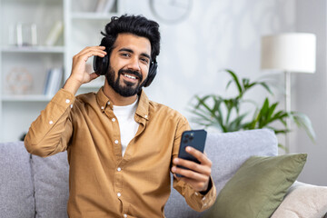 Satisfied Arab man with a beard in headphones sitting on the sofa listening to music, holding a...