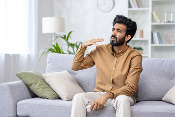 Young Indian man with beard sits in the living room in summer, fanning his hand, suffering from the heat, holding the air conditioner remote control, unpleasant temperature in the room, overheating.