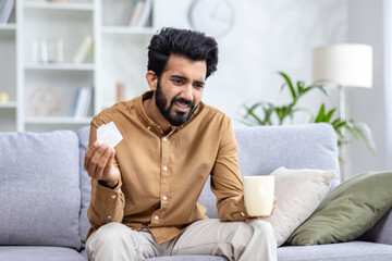 Sick Hindu man sitting on sofa at home, holding cup in hand, drinking tea, holding napkin, viral diseases, runny nose and cough, health concept.
